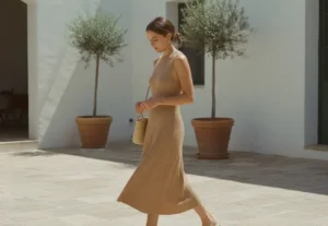 Minimalist lifestyle photography of a woman in a beige ribbed midi dress and tan sandals in a sunny courtyard.