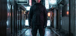 A full-body shot of a man standing in a wet, dark urban alleyway lit by neon signs at night. He is wearing a black techwear outfit consisting of a multi-pocket jacket, cargo pants, and gloves.