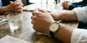 A close-up candid shot of a man's hands resting on a rustic wooden table during a meeting or dinner. A classic stainless steel automatic watch with a black leather strap is prominent on his wrist.