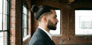 A side profile view of a bearded man in a tweed jacket. His long hair is pulled back tightly into a neat man bun, contrasting with a sharp taper fade on the sides and back of his head.