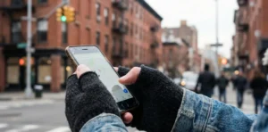 A close-up of hands wearing black knit fingerless gloves holding a smartphone with a cracked screen displaying a map app, against a blurred urban brick background.