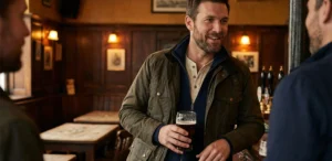A man in a waxed jacket and knit cardigan holds a pint of beer and talks in a traditional pub.