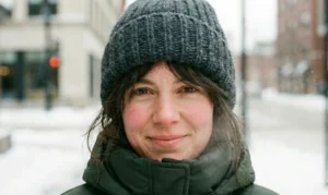 A portrait photograph of a woman's face on a winter day. She wears a chunky charcoal grey knit beanie and a dark green jacket collar is visible. Her cheeks are rosy, and she smiles gently at the camera with snowy buildings in the background.