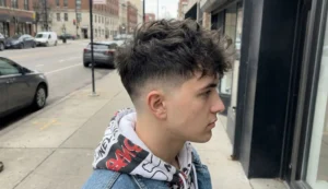 A portrait of a young man with a messy, layered, medium-length shag or "wolf cut" hairstyle, wearing a denim jacket in a cluttered studio space.