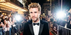 A paparazzi-style photograph of actor Liam Hemsworth featuring textured blonde hair. He is wearing a black tuxedo and bow tie, walking a red carpet surrounded by intense camera flashes and a crowd of onlookers. A sign for "FESTIVAL DE CANNES" is visible in the background.