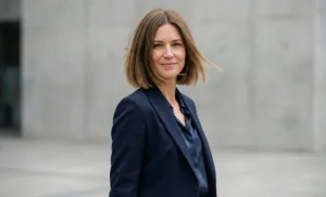 Portrait of a professional woman with a chin-length blunt brown bob and a side part, wearing a navy blazer against a concrete building background.