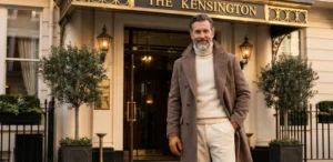 A well-dressed older man in a brown wool coat and cream turtleneck smiles in front of "The Kensington" luxury hotel entrance.