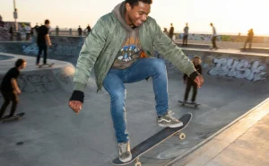 A young Black man smiles as he skateboards in a skatepark during sunset.