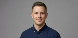 A professional studio portrait of a man smiling against a grey background, wearing a blue shirt and a modern crew cut that is textured on top and neatly tapered on the sides.