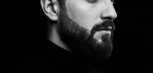 A high-contrast, black and white profile portrait of a man showing a very sharply shaped full beard with precise cheek lines.