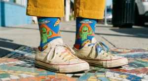A low-angle shot of a person's ankles on a colorful mosaic sidewalk, wearing bright blue socks with cartoon cacti, UFOs, and pizza, cropped yellow corduroy pants, and worn white sneakers with rainbow laces.