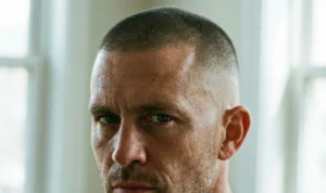 An intense, extreme close-up portrait of a man's face. He has a disciplined military-style buzz cut with a precise high skin fade on the sides. Sweat and stubble are visible on his face.