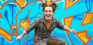 A creative shot of a young man with double twisted space buns, wearing a patterned jumpsuit and laughing while jumping in front of a bright, colorful graffiti wall.