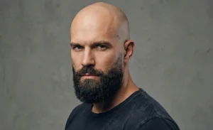 A portrait of a completely bald man with a thick, dark beard that fades into the scalp near the ears, against a grey studio background.