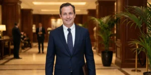 A photograph of a man in a dark navy suit and tie, holding a leather portfolio and smiling slightly, standing in a warmly lit, luxurious hotel lobby with wood paneling and plants.