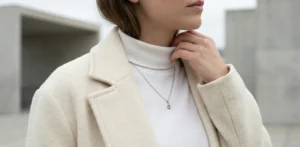 A close-up detail shot of a woman wearing a ribbed white turtleneck sweater under a textured cream wool coat, accessorized with a delicate silver pendant necklace.