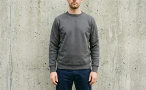 A portrait of a man standing confidently against a textured concrete wall, wearing a plain, high-quality dark grey crewneck sweatshirt and dark blue chinos.