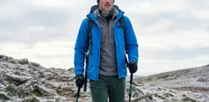 A man in a blue technical jacket and hiking gear walks on a snowy, rocky trail with trekking poles.