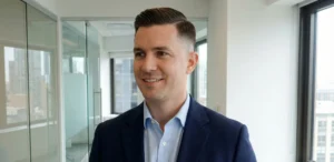 A smiling professional man standing in a modern office with glass walls. He is wearing a navy blazer and has a neat side-part haircut with a subtle, balanced fade on the sides.
