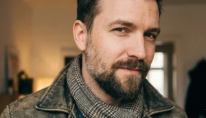 Portrait of a man with a short, textured full beard and a slight smirk, wearing a distressed brown leather jacket and a patterned scarf indoors.