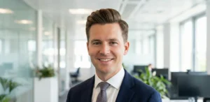 A professional headshot of a smiling man with neat, styled brown hair wearing a blue suit and patterned tie in a bright corporate office setting.