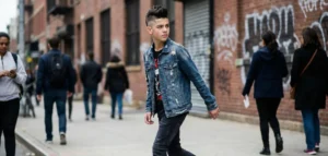 Street style photo of a teenage boy with a faux hawk fade haircut walking on a city sidewalk, wearing a distressed denim jacket.