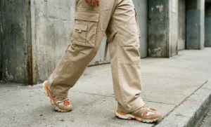 A low-angle shot focusing on a person's legs walking on concrete, wearing wide-leg beige cargo pants with straps and chunky tan and orange sneakers.
