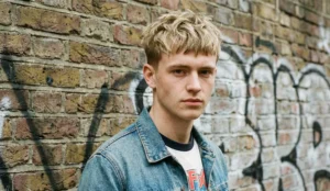 A portrait of a young man with a messy, textured blonde French crop haircut and fringe, standing in front of a graffiti-covered brick wall.