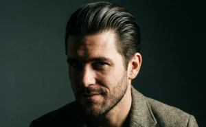 A close-up, moody portrait of a man with dark, slicked-back hair and stubble, looking directly at the camera.