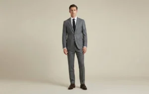 A full-body studio photograph of a man standing against a neutral beige background, demonstrating a perfectly fitted grey suit, white shirt, black tie, and brown monk strap shoes.