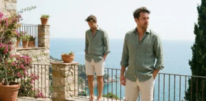 Two men standing on a sunlit stone balcony overlooking the Mediterranean Sea. Both are wearing sage green linen shirts with rolled sleeves and stone-colored linen shorts. Bright daylight illuminates the scene, with pink flowers and terracotta pots nearby.
