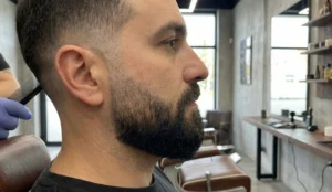 A side-profile photograph showing a man's fresh haircut and tapered beard fade in a modern barbershop, with a barber's hand holding a comb near his ear.
