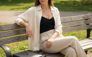 A woman sitting relaxedly on a wooden park bench on a sunny day. She wears an open cream silk shirt over a black bodysuit, beige linen trousers, and layered gold necklaces. A laptop in a case rests on the bench beside her.