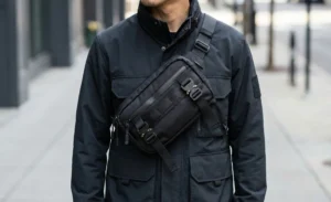 A close-up detail shot of a man's torso. He is wearing a black technical crossbody bag with straps and buckles diagonally across his chest, over a black utility jacket.