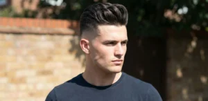 Profile view of a man with a sharp high fade pompadour haircut, standing outdoors against a sunlit brick wall background.