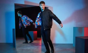 A conceptual fashion shot in a studio with creative lighting, showing a male model in a reversible bomber jacket; one side is plain black, and a mirror reflection shows the other side with a colorful geometric pattern.