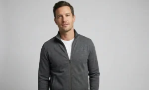 A studio portrait of a man in a simple, high-quality grey zip-up jacket and a white t-shirt, standing against a plain light grey background