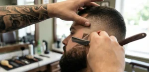 A close-up shot from behind the shoulder of a barber using a straight razor to etch a design into the fade of a client's beard.
