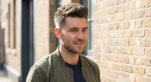 A candid portrait photograph of a man with a textured quiff hairstyle, wearing an olive green bomber jacket over a t-shirt, standing outdoors in front of a blurred brick building.