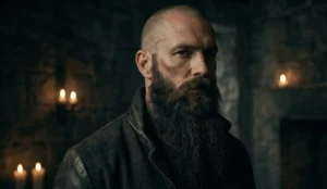 A portrait of a man in a "modern Viking" style with a shaved head and a long, flowing beard against a dark, stone background.