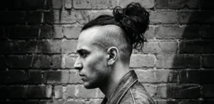 A high-contrast black and white profile photograph of a man with a large, messy man bun and shaved sides. He is wearing a leather jacket and an earring, standing against a brick wall.