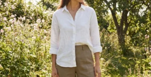 A woman wearing a white linen button-up shirt and brown linen trousers stands in a sun-drenched garden filled with wildflowers.