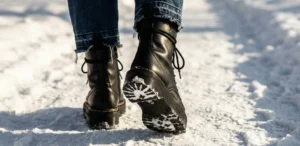 A low-angle, close-up view from behind of black leather platform combat boots with thick, rugged soles walking away on a snowy path.