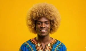 A portrait of a smiling Black man with a large, golden-blonde dyed afro hairstyle, wearing a colorful patterned shirt against a bright yellow background.