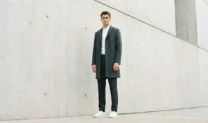 A man in a charcoal coat and white sneakers poses minimalist-style against a large, textured concrete wall.
