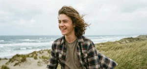 A candid outdoor shot of a laughing teenage boy walking through coastal dunes on a windy day. His medium-length brown hair is blowing backward in a relaxed "bro flow" style, and he wears a plaid flannel shirt.