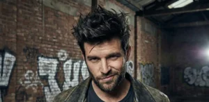 A close-up of a man with messy, spiky dark hair and a beard, wearing a worn leather jacket and giving a smirk against a brick wall covered in graffiti.