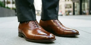 A low-angle close-up shot focusing on a pair of impeccably polished brown leather cap-toe oxford shoes worn by a man standing on a concrete sidewalk. The cuffs of dark grey suit trousers are visible just above the shoes.