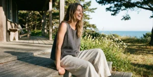 A woman with wavy hair laughs with her head tilted back, sitting on wooden steps outdoors. She is wearing a loose-fitting charcoal grey sleeveless linen top and light beige wide-leg linen pants. In the background are a rustic building, wildflowers, and a view of the ocean under a sunny sky.