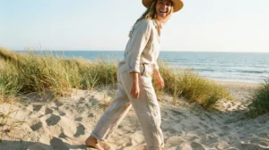 A woman wearing a wide-brimmed straw hat, a long-sleeved oatmeal-colored linen shirt, and matching loose linen pants laughs as she walks barefoot on sandy dunes by the beach. The texture of the linen fabric is prominent in the sunlight. Tall dune grasses and the ocean are in the background.